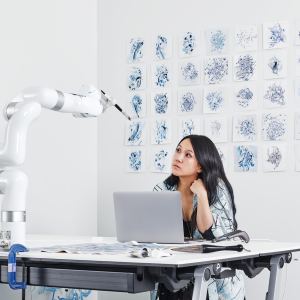 A woman with long dark hair sits at a desk with a laptop, looking thoughtfully at a white robotic arm holding a paintbrush. The desk has art supplies, including brushes and tubes of paint. Behind her, a wall is covered with multiple small abstract blue ink or watercolor paintings arranged in a grid pattern. The setting appears to be a modern, minimalist studio or workspace.
