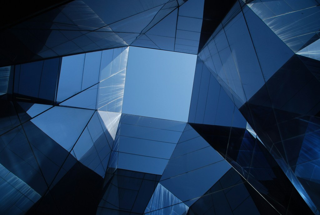 View looking up through a geometric arrangement of reflective blue glass panels forming a square opening to the clear blue sky above. The panels create angular shapes and reflections, giving a modern and abstract architectural appearance.