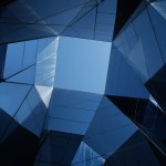 View looking up through a geometric arrangement of reflective blue glass panels forming a square opening to the clear blue sky above. The panels create angular shapes and reflections, giving a modern and abstract architectural appearance.