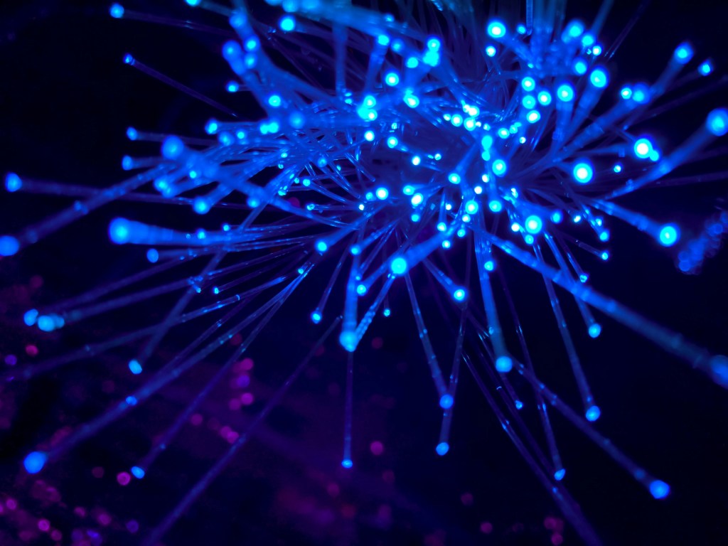 Blue fiber optic strands glowing against a dark background with some purple light spots. The strands radiate outward with bright blue tips.