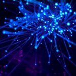 Blue fiber optic strands glowing against a dark background with some purple light spots. The strands radiate outward with bright blue tips.