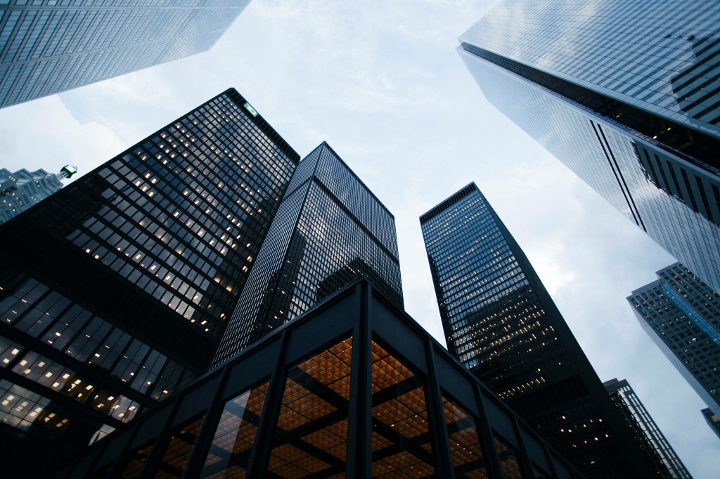 Tall modern skyscrapers with glass facades viewed from below against a cloudy sky, featuring illuminated office windows and a dark building corner with warm interior lighting.